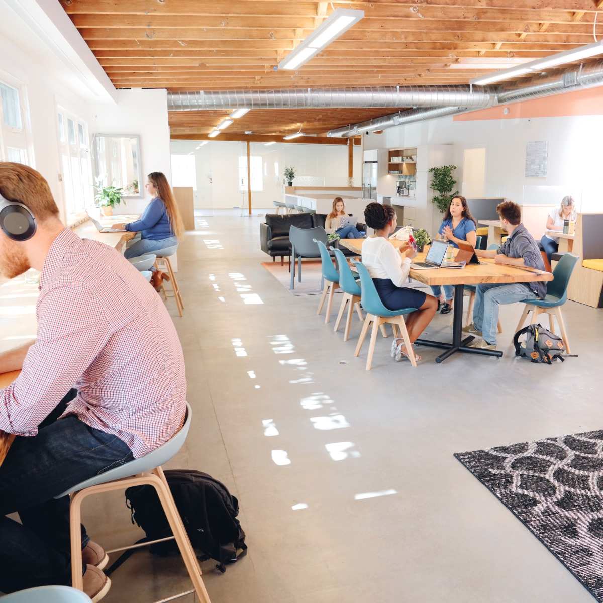 A modern workspace features people collaborating at tables, with a mix of seating arrangements and natural light flooding the area.