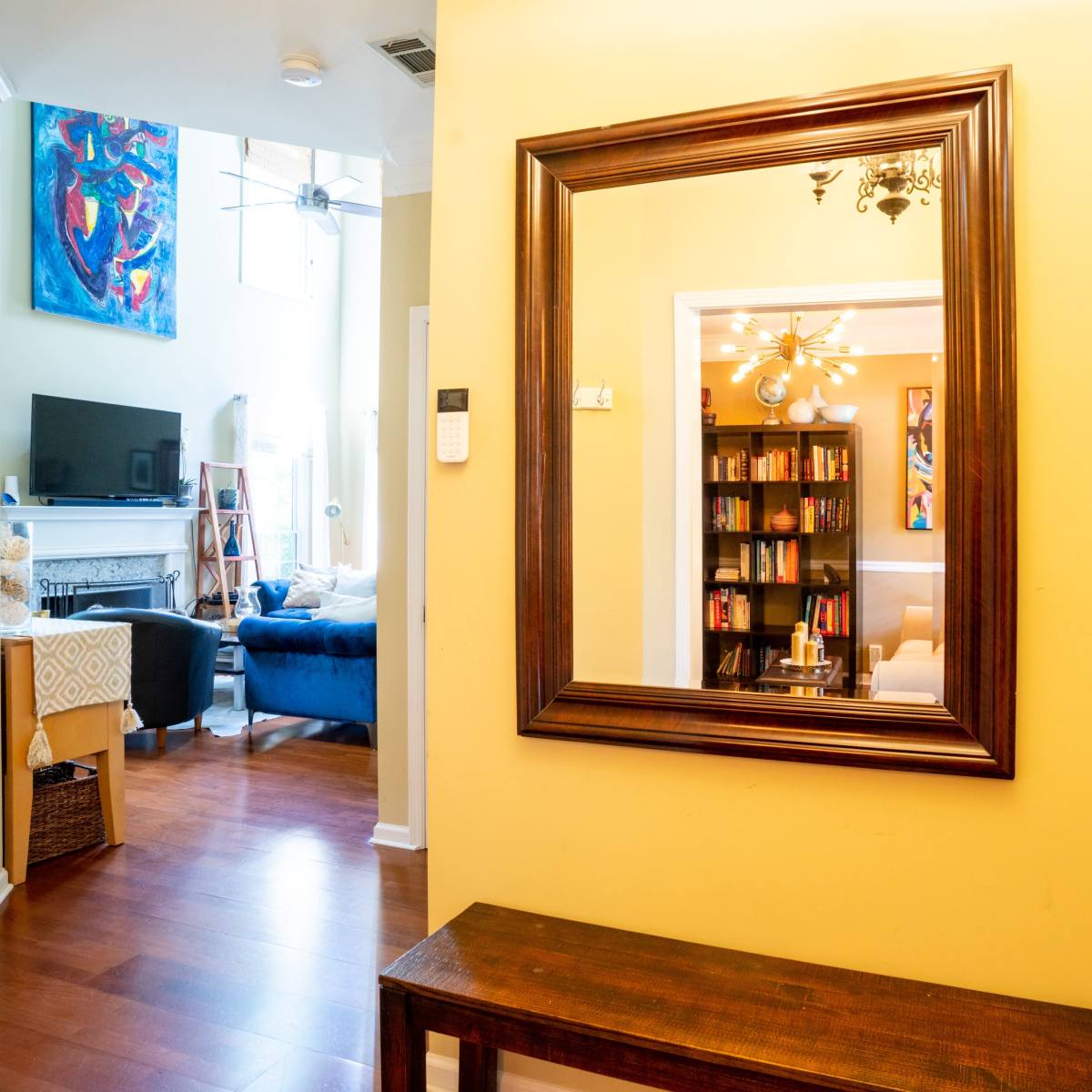 A hallway with a large mirror on the wall, a wooden bench in front, and a view into a living room featuring bookshelves and a TV in the background.