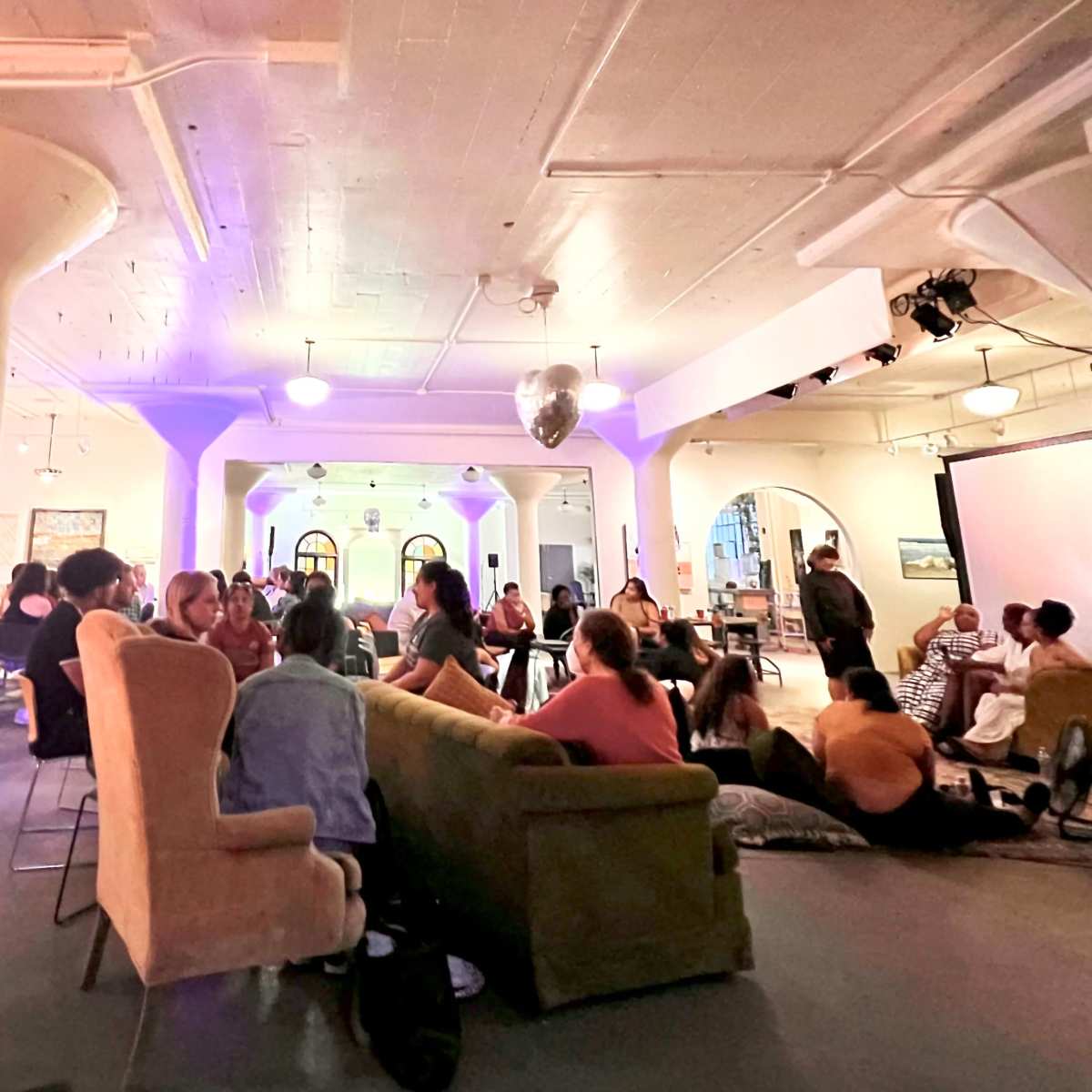 A group of people gather in a spacious, well-lit room with colorful lighting and various seating arrangements for an event.