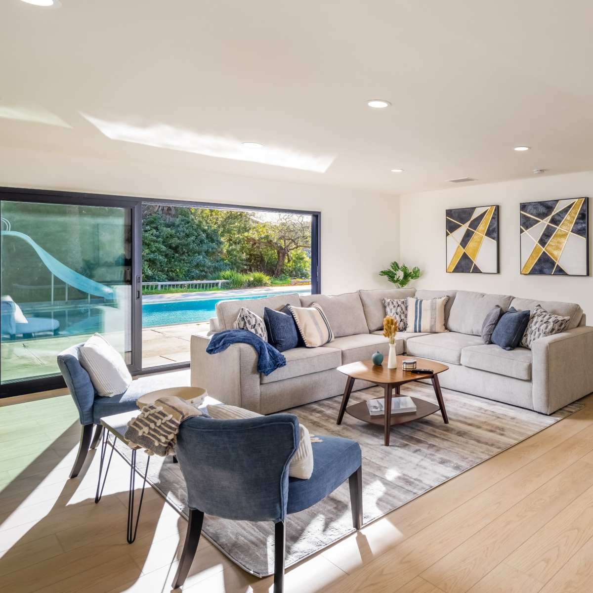 A spacious living room features a light-colored sectional sofa, two blue chairs, and large windows overlooking a pool area.