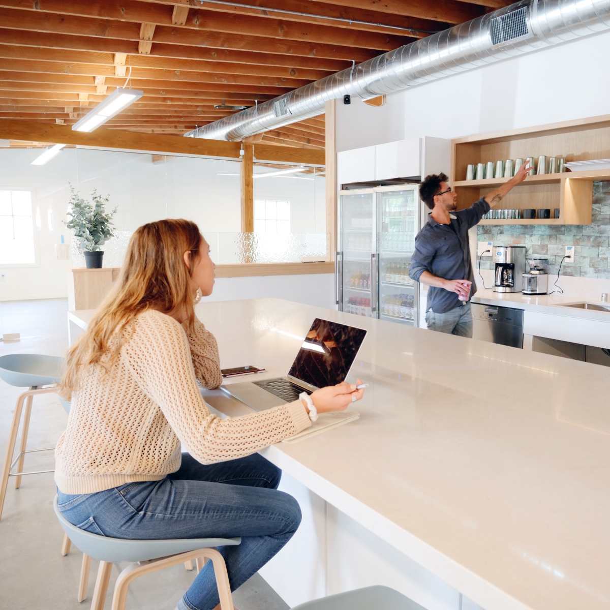 A man arranges cups on a shelf while a woman stands nearby, and another woman sits at a table with a laptop in a modern kitchen space.