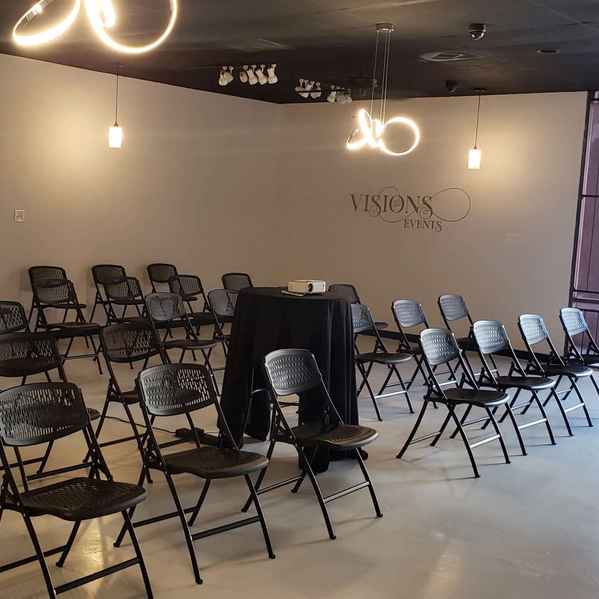 A spacious room with rows of black folding chairs arranged facing a central table, illuminated by pendant lights.