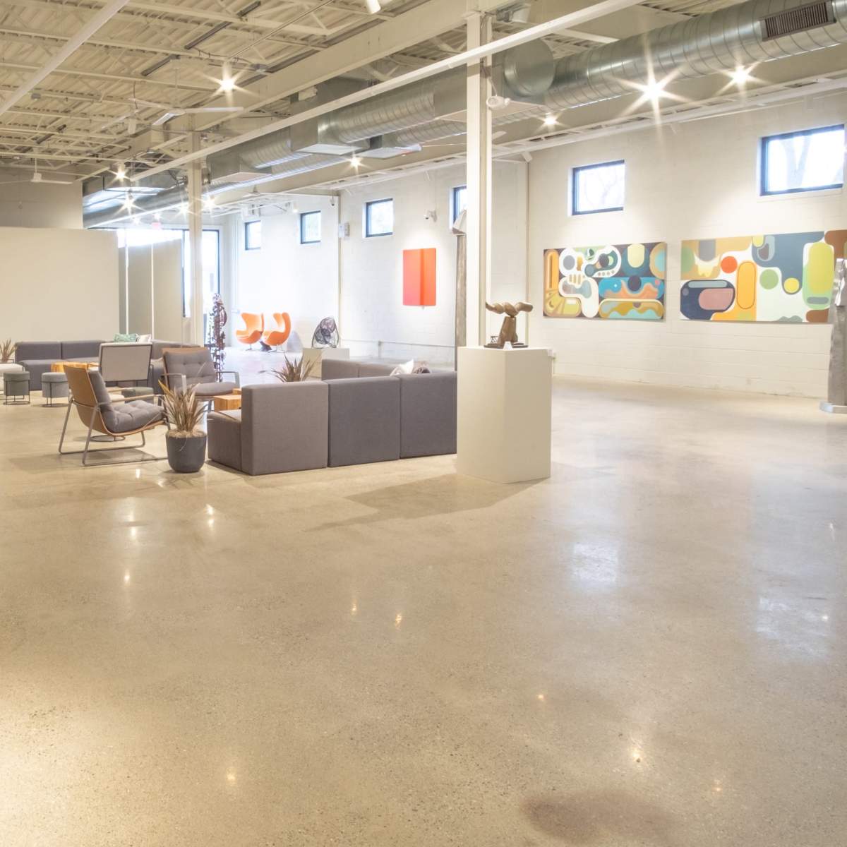 An art gallery featuring various colorful paintings and sculptures arranged in an open, well-lit space with concrete flooring and seating areas.