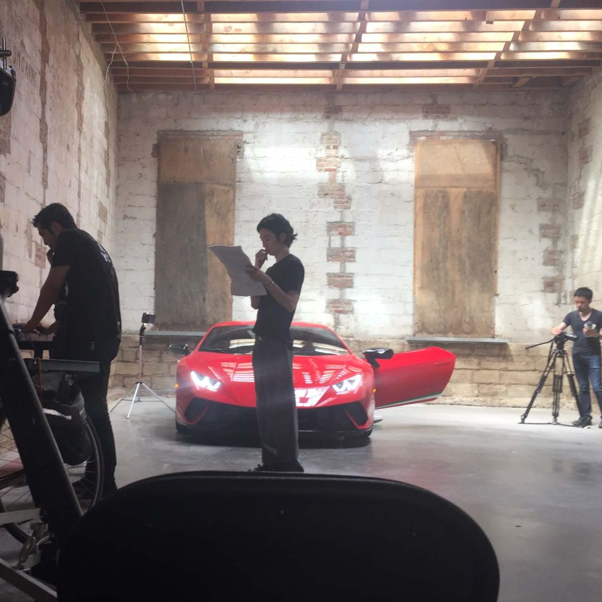 Three individuals are engaged in different activities in a well-lit garage, with a red sports car prominently featured in the background.