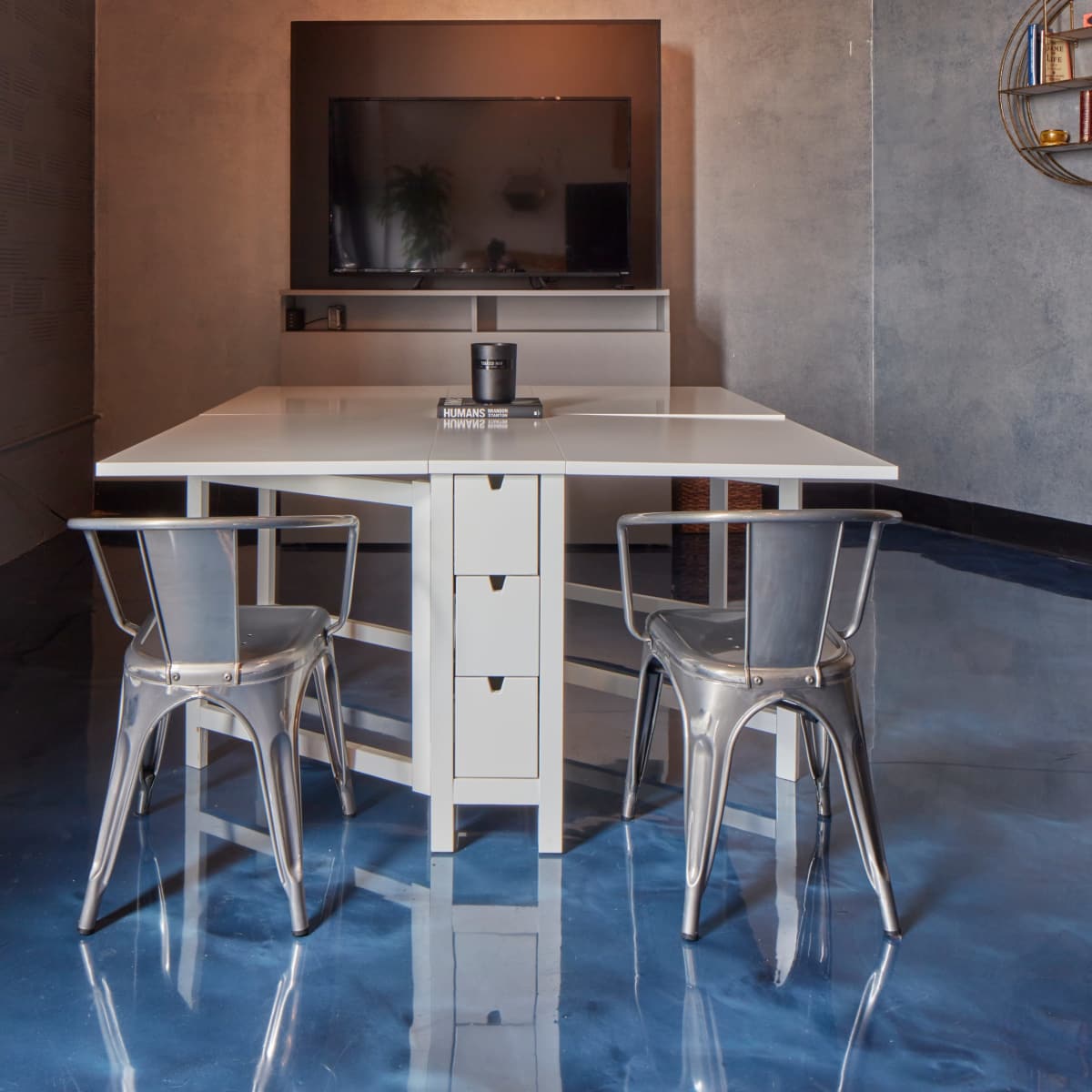 A white table with metal chairs is positioned in a room featuring a television mounted on the wall and a glossy blue floor.
