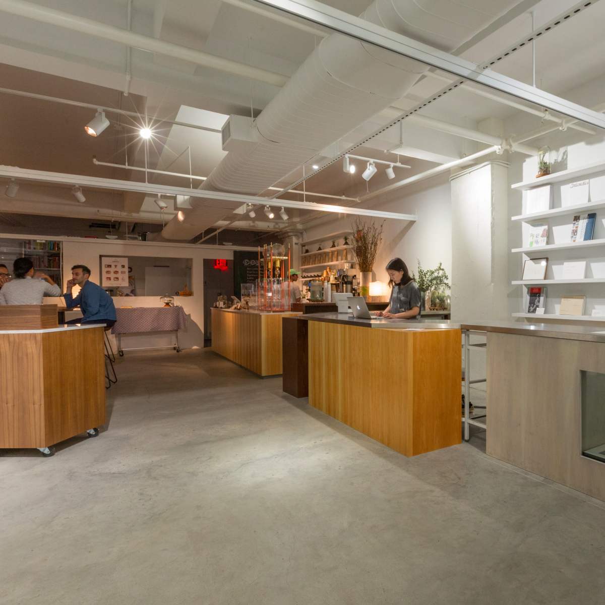 A modern interior of a shop or cafe with wooden counter spaces, shelves displaying various items, and a few people engaging at the counters.