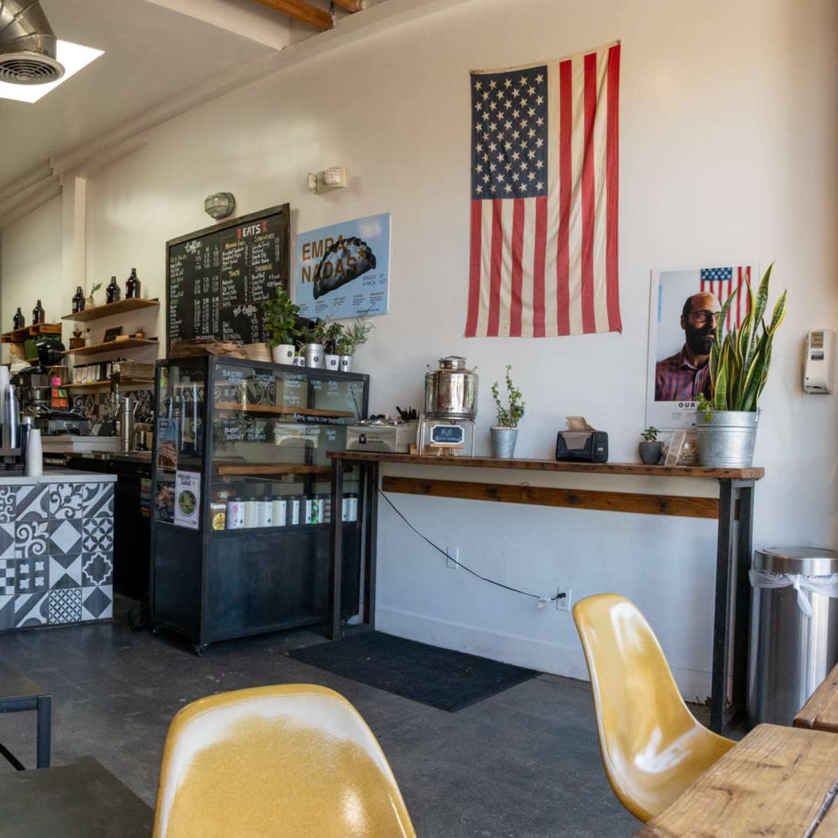 A coffee shop interior with brick walls, a counter displaying various beverages, seating areas, and an American flag on the wall.
