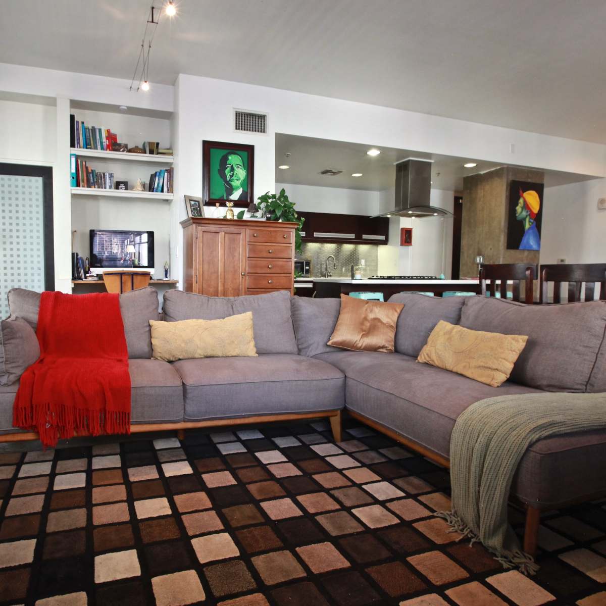 A contemporary living room featuring a grey sectional sofa with a red throw, a patterned area rug, and a kitchen visible in the background.