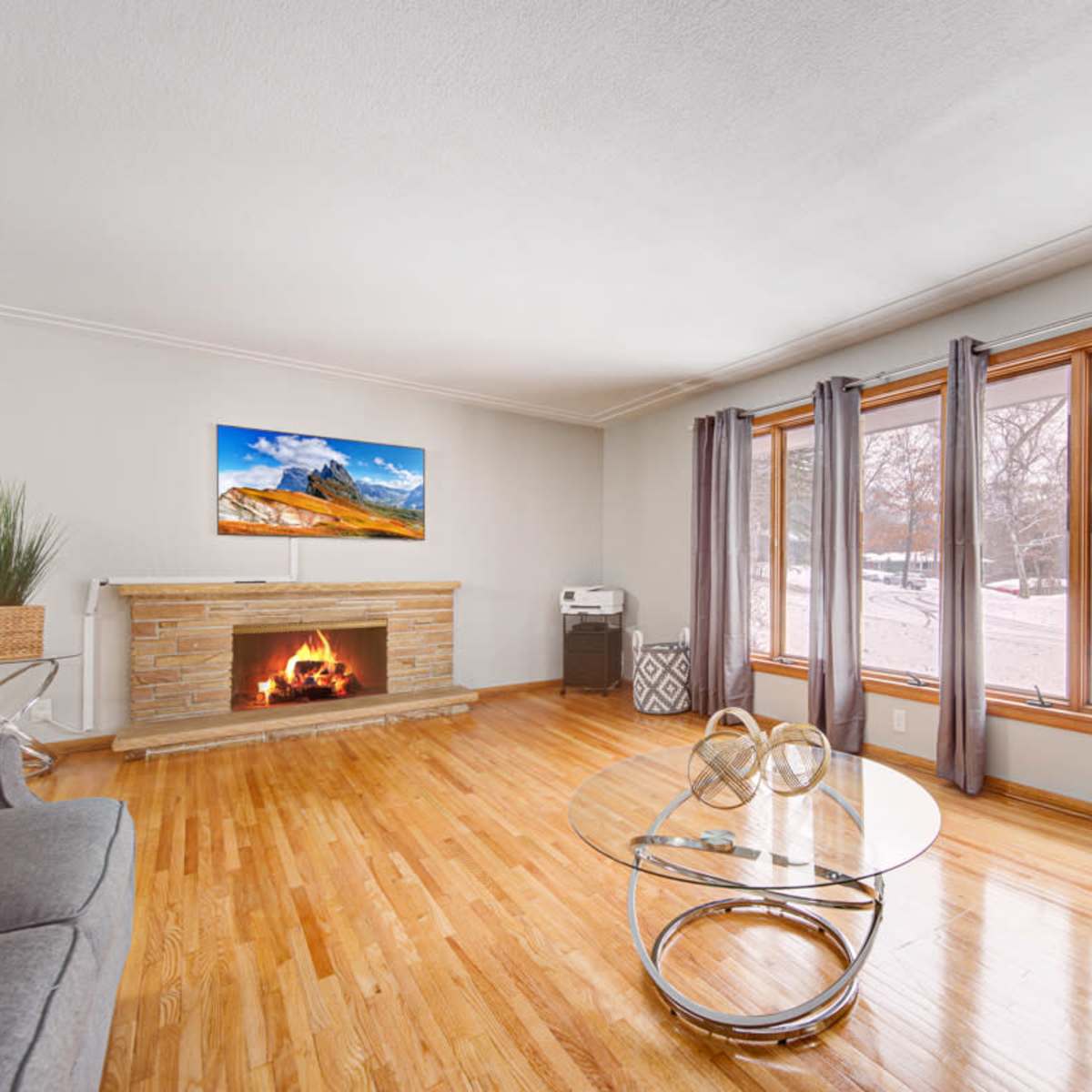 The living room features hardwood floors, a stone fireplace with a television above it, large windows with views of a snowy outdoor scene, and a round glass coffee table.