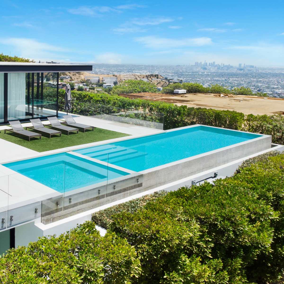 A modern house features a sleek swimming pool and a landscaped terrace with lounge chairs, overlooking a city skyline.