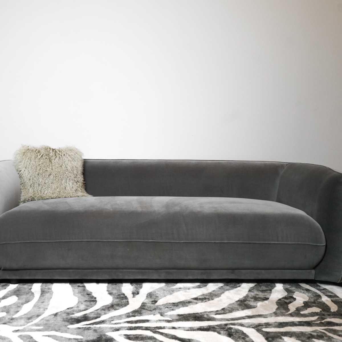 A gray upholstered sofa with rounded arms and a textured pillow sits on a zebra-striped rug against a plain wall.