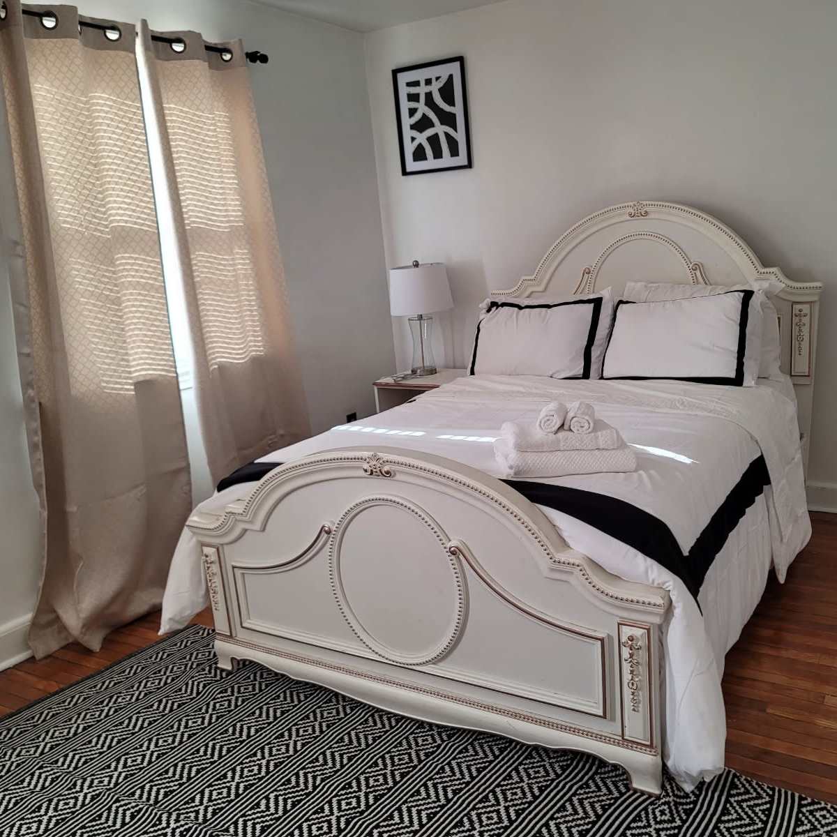 A neatly arranged bedroom features a bed with white linens and black accents, a lamp on a nightstand, and curtains letting in natural light.