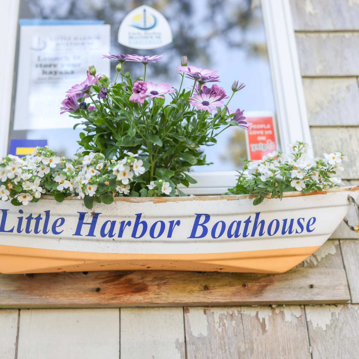 A small, decorative boat filled with colorful flowers is displayed outside a window, labeled "Little Harbor Boathouse."