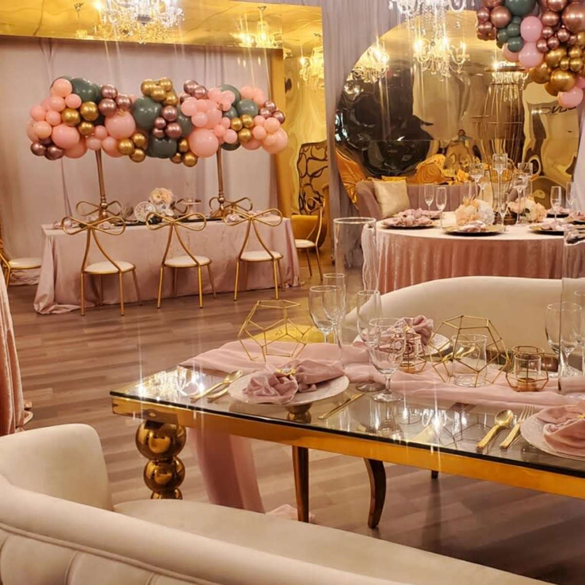 A decorated banquet hall featuring a dining table set with gold accents, pink napkins, and a backdrop of colorful balloons.