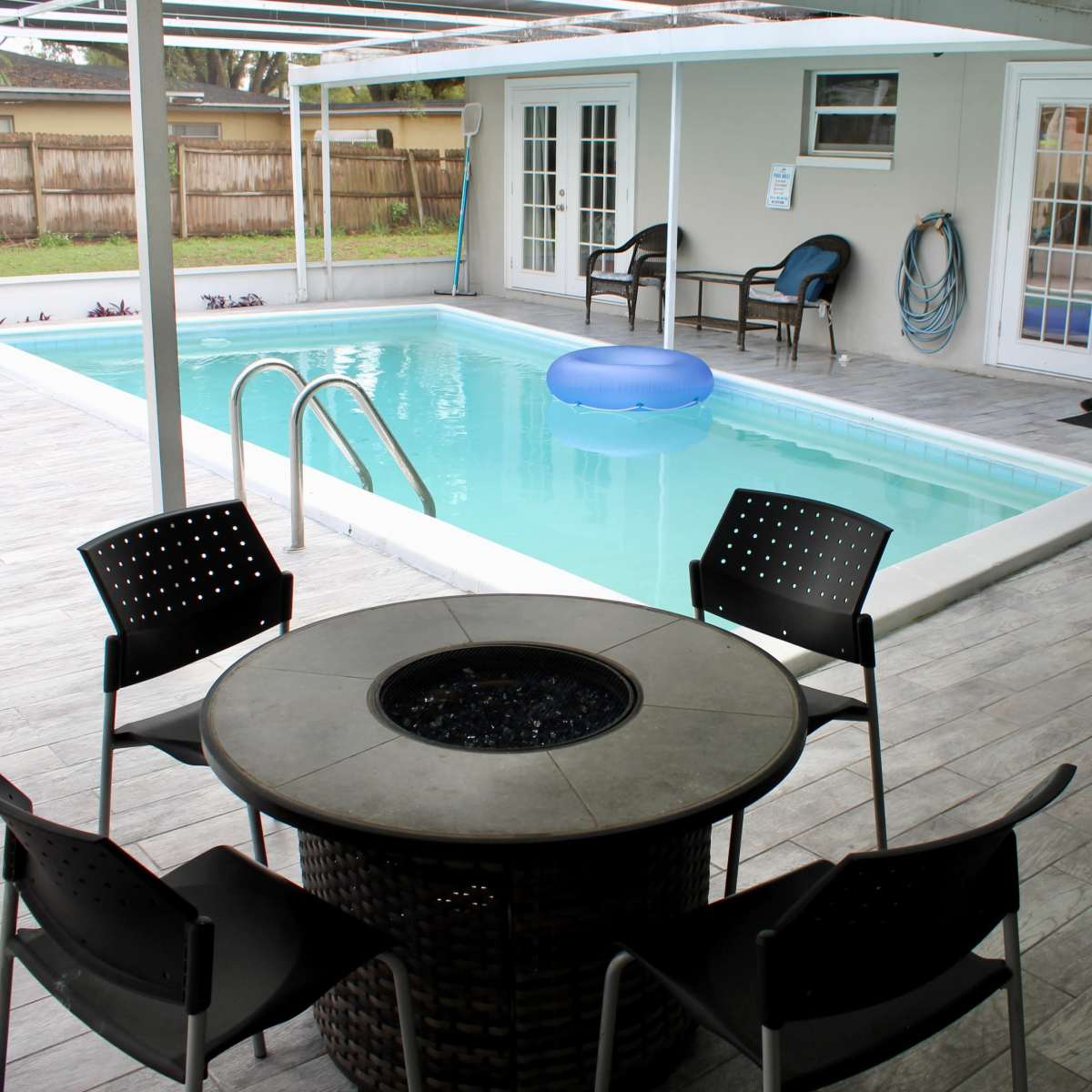 A swimming pool with a blue float, surrounded by a covered patio area featuring a circular fire pit table and black chairs.