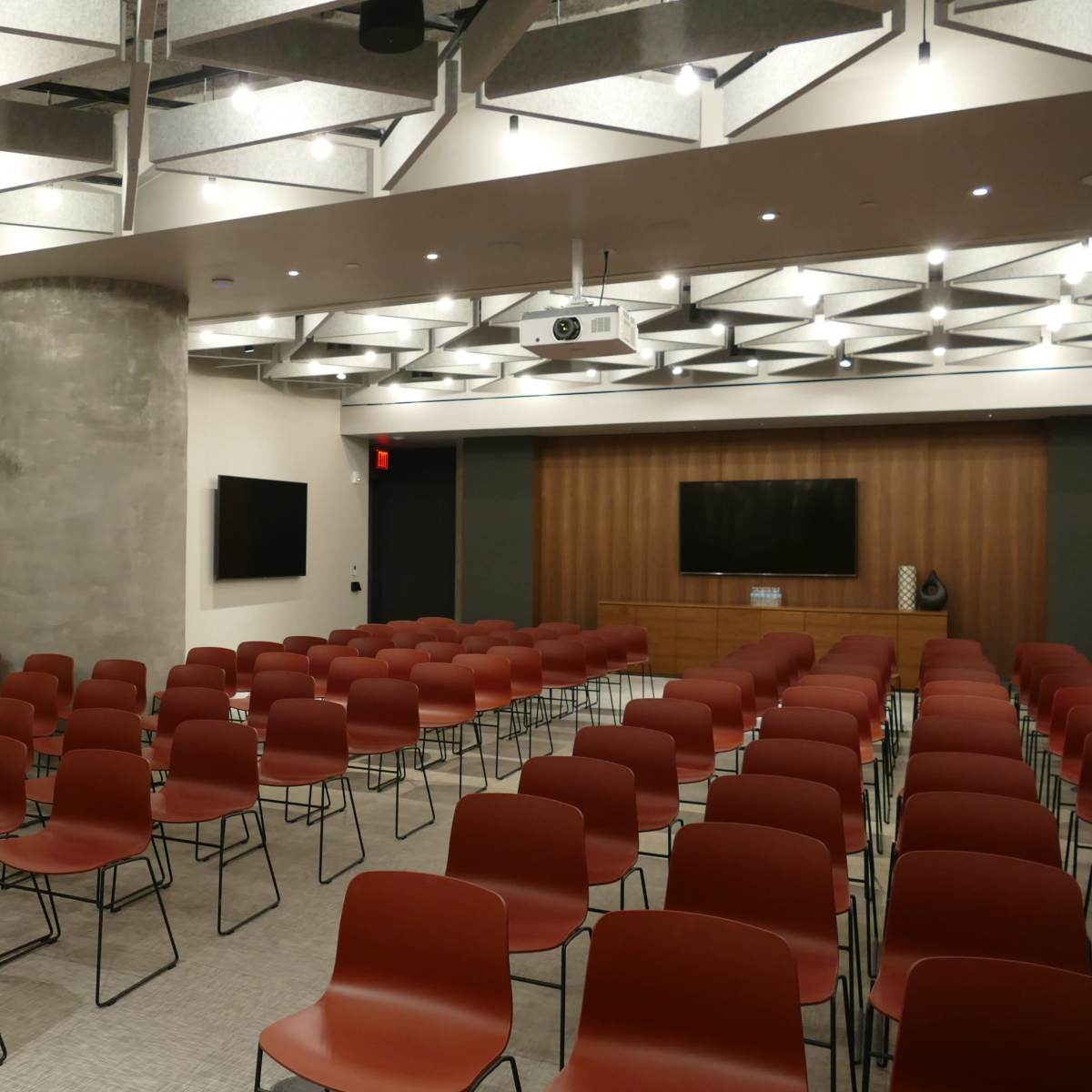 A modern conference room set up with multiple rows of red chairs facing a wooden presentation area, featuring two large screens on the walls.
