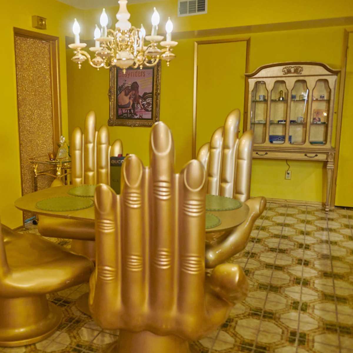 The room features a bright yellow interior with a dining table surrounded by gold hand-shaped chairs and a chandelier overhead.