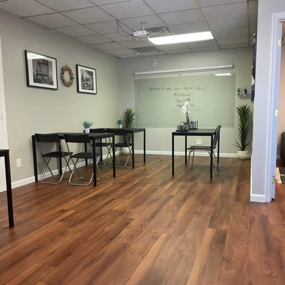 A spacious room with wooden flooring, featuring several black tables and chairs, along with wall decorations and plants.