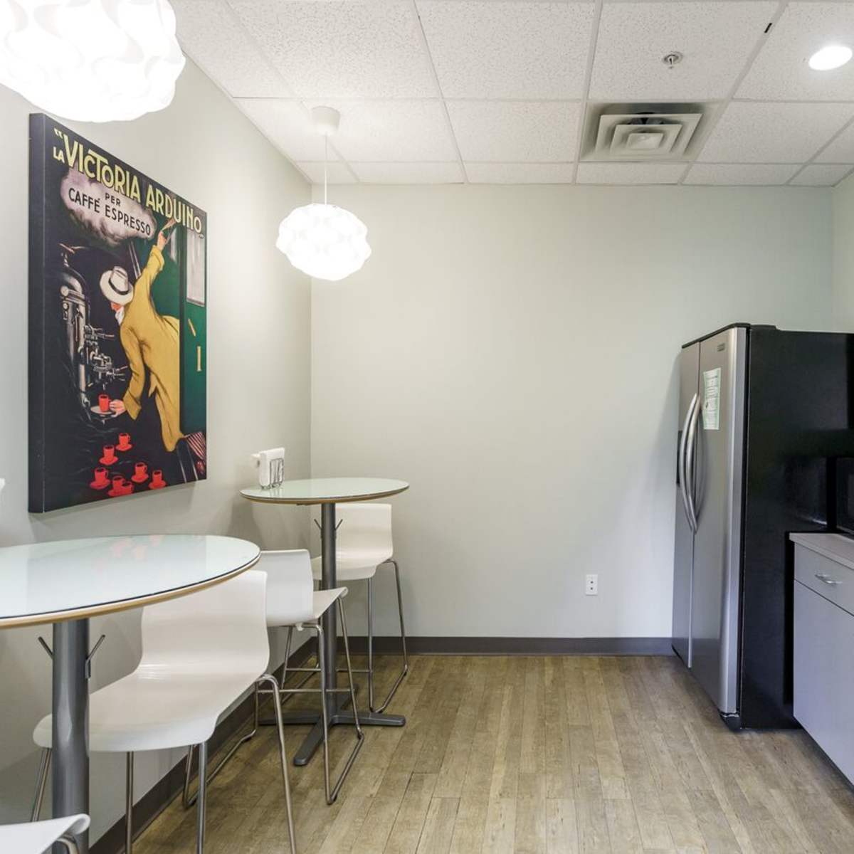 A modern break room with a round table, four high-backed stools, a refrigerator, a microwave, and a wall-mounted artwork.