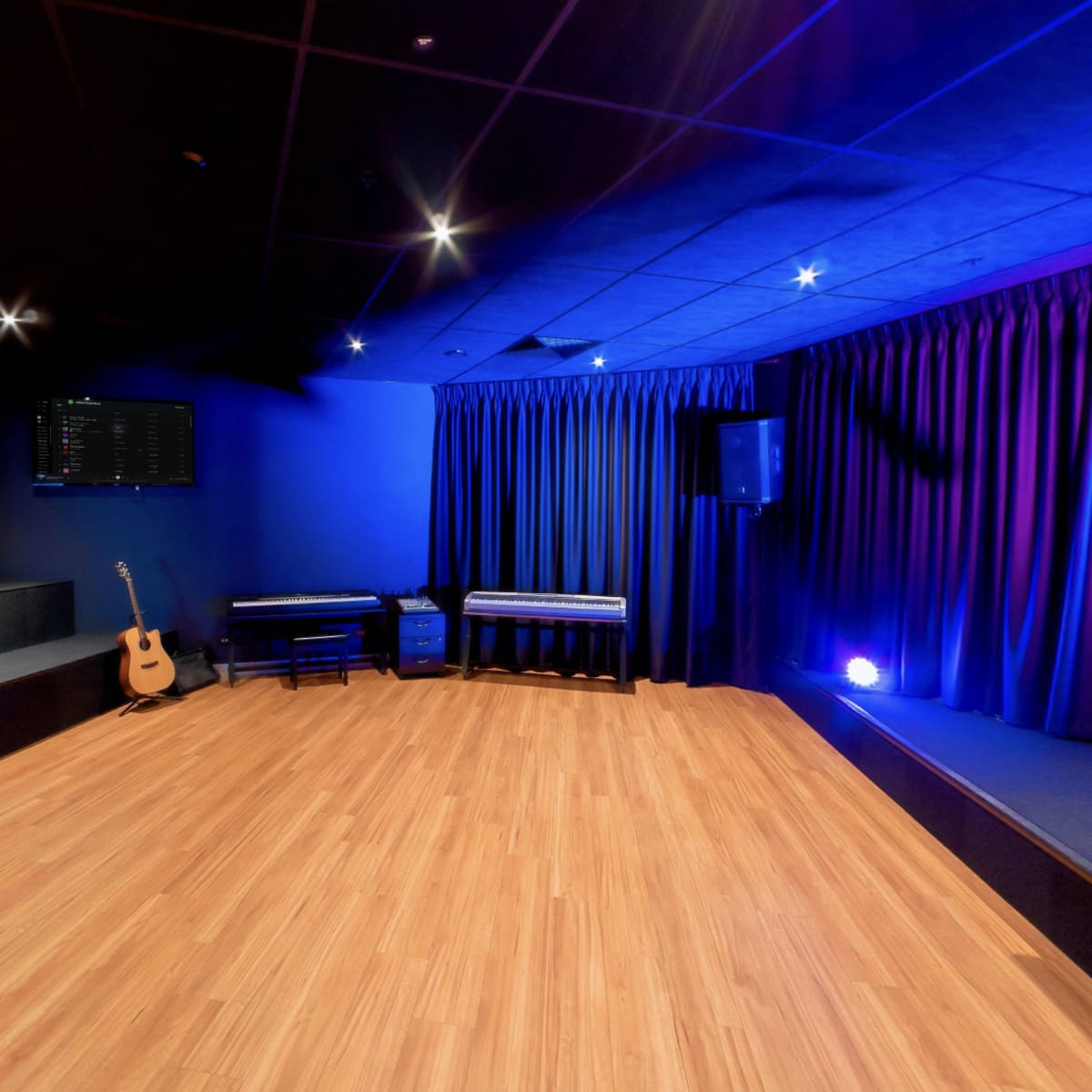 A performance space with a wooden floor, a stage area, a microphone stand, and sound equipment visible through a glass window.