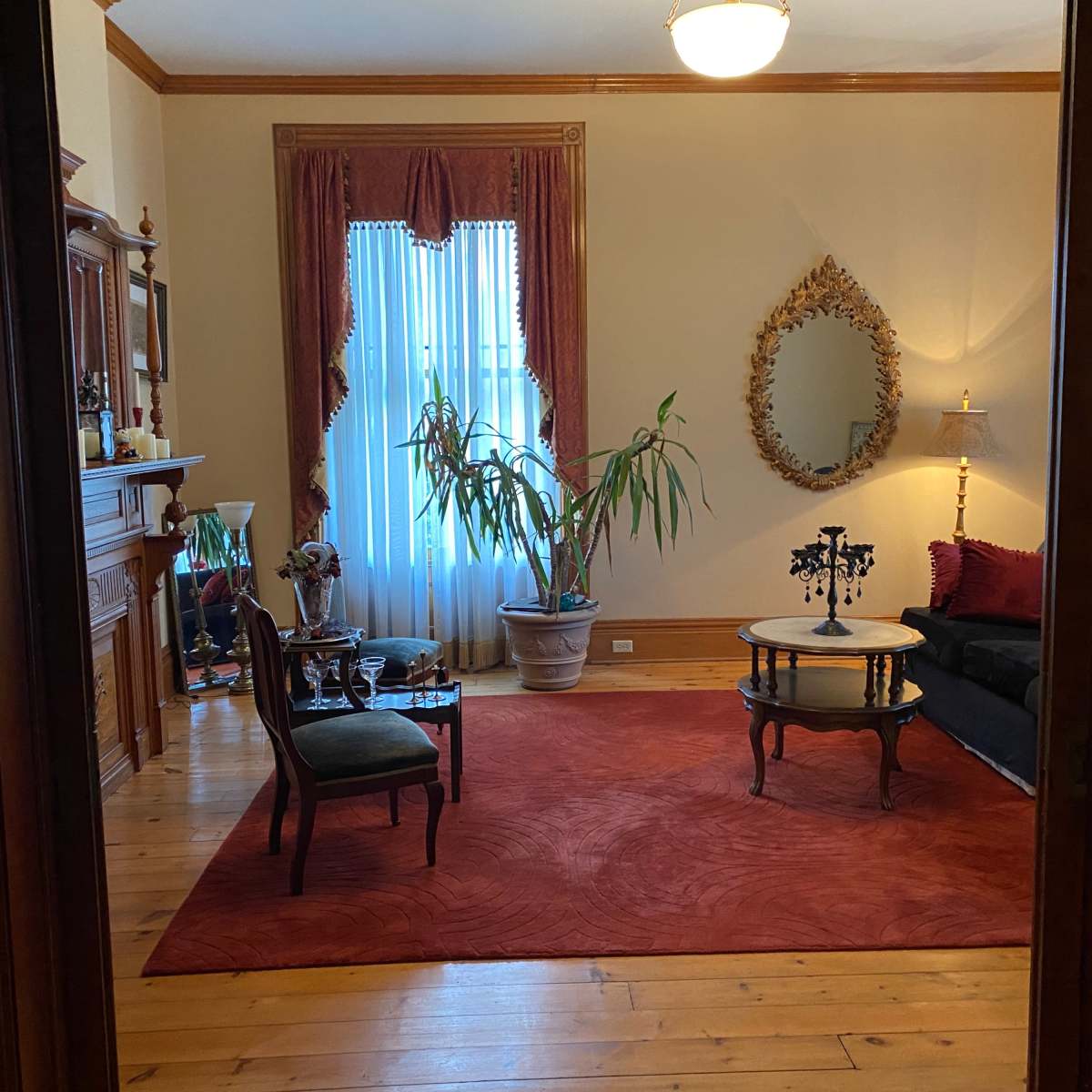 A living room with a large rug, a black sofa, a decorative mirror, and a plant near a window with curtains.