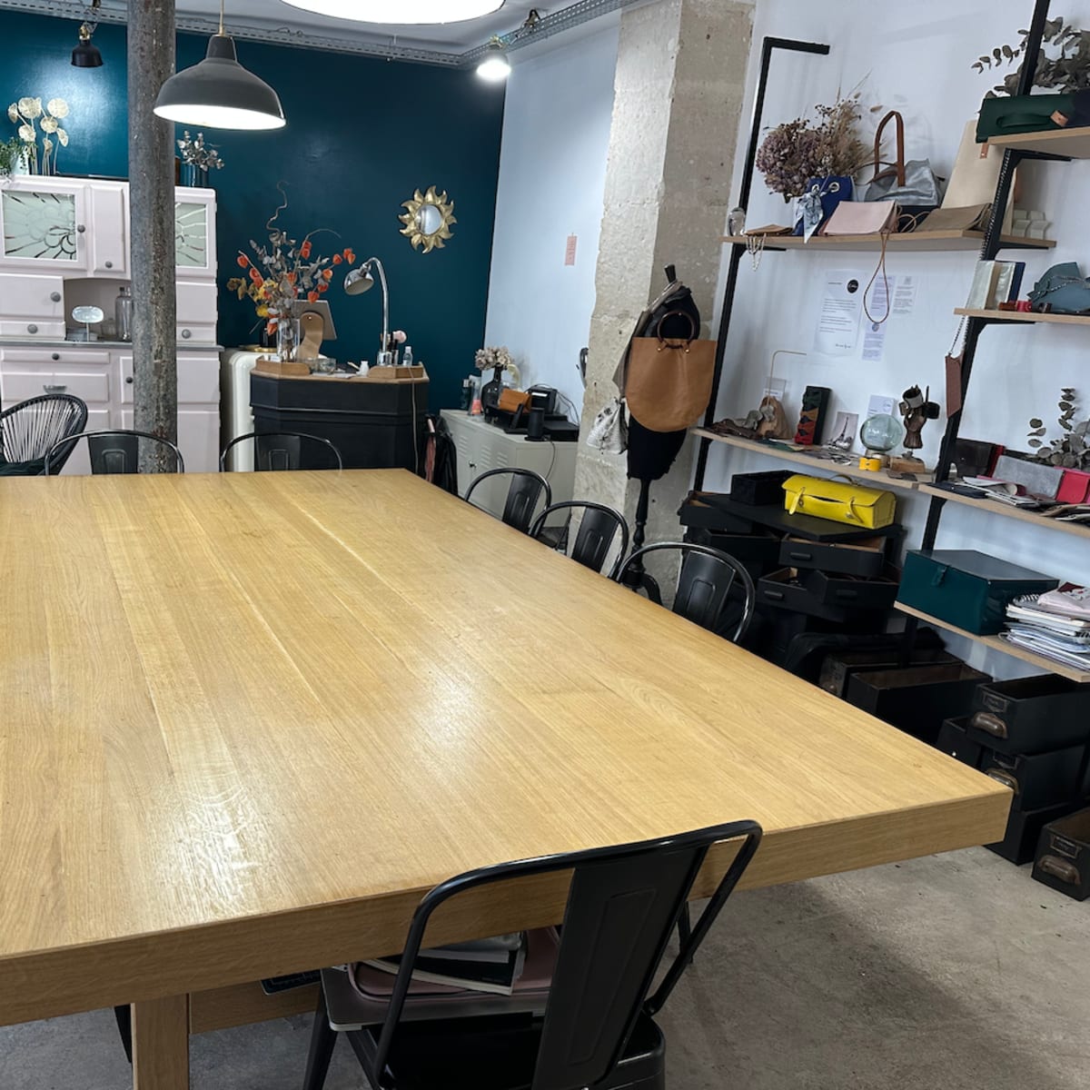 A large wooden table surrounded by black chairs in a well-decorated workspace with a green accent wall and various items on shelves.