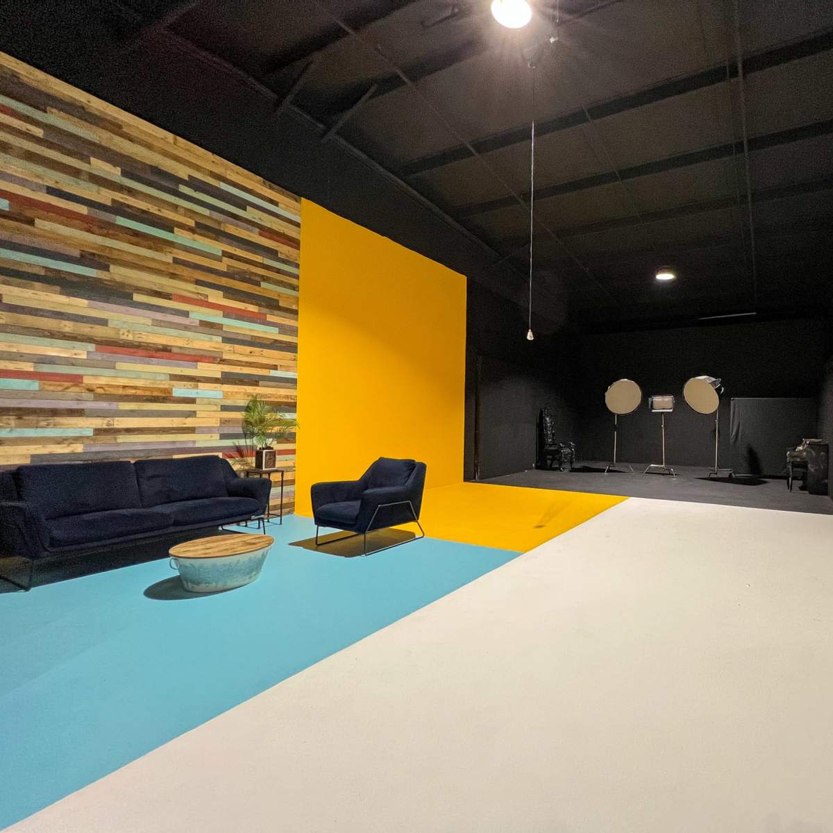 A spacious studio with a textured wooden accent wall, a bright yellow section, a light blue area, and a white backdrop, featuring a sofa and standing equipment.