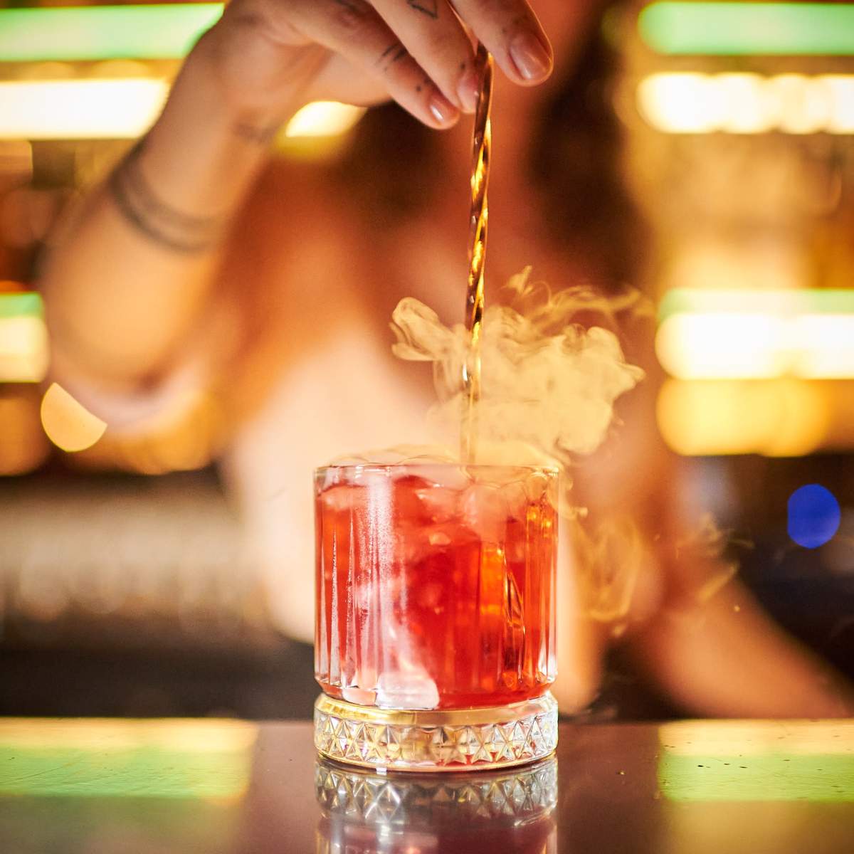 A bartender stirs a cocktail in a glass with ice, while vapor rises from the drink.