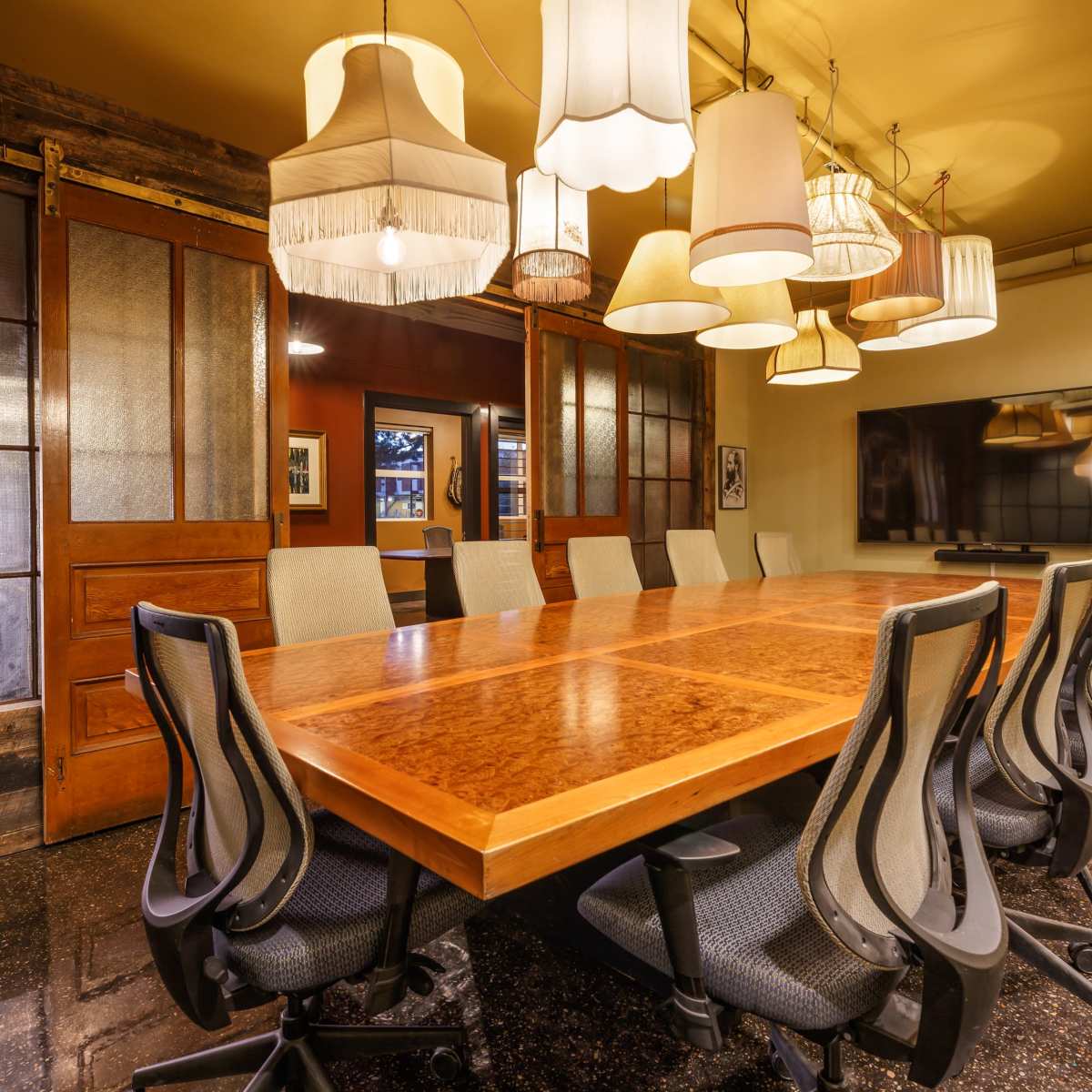 A long wooden conference table surrounded by ergonomic chairs is illuminated by several pendant lamps hanging from the ceiling.