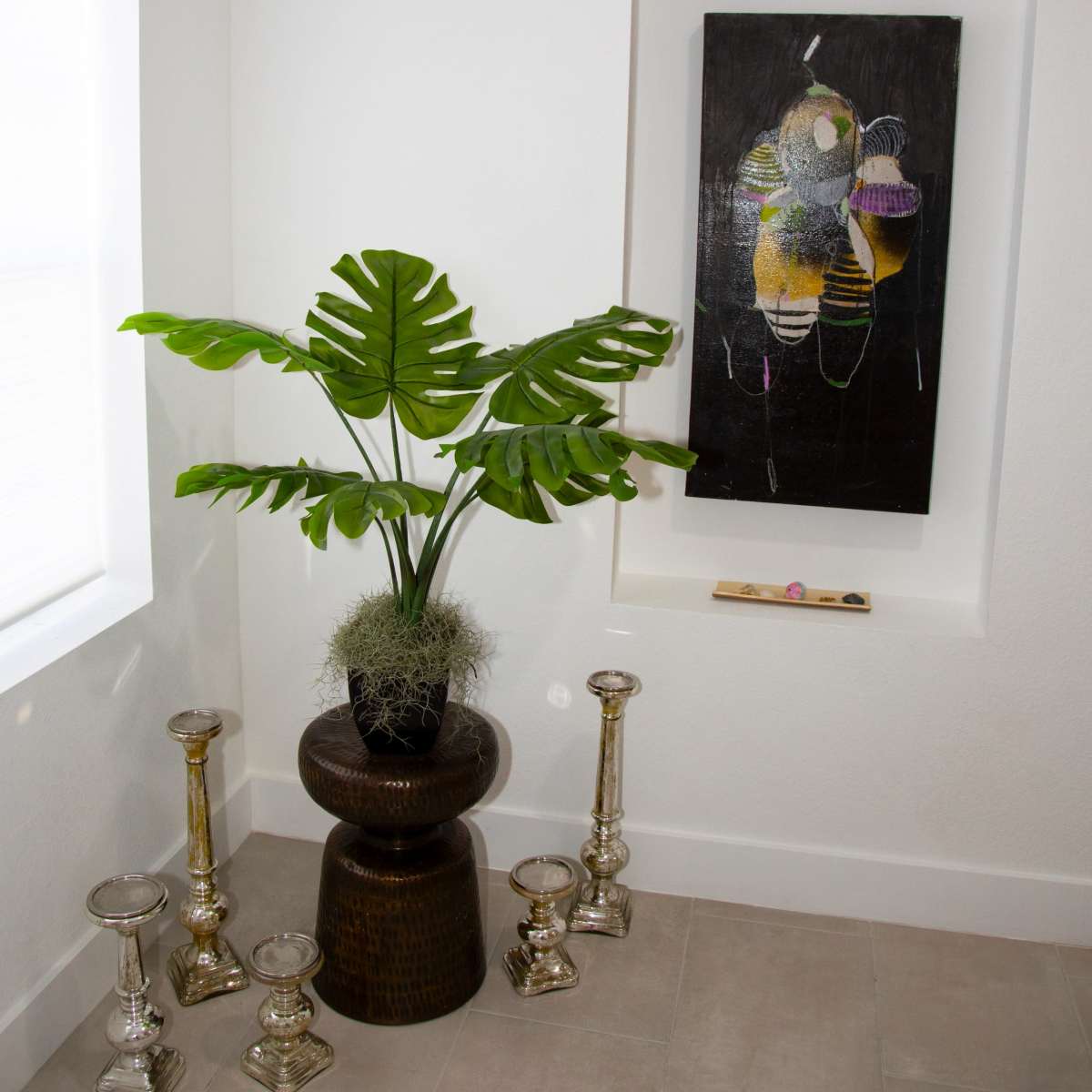A potted plant with large leaves stands beside decorative candle holders and a framed abstract painting in a bright corner of a room.