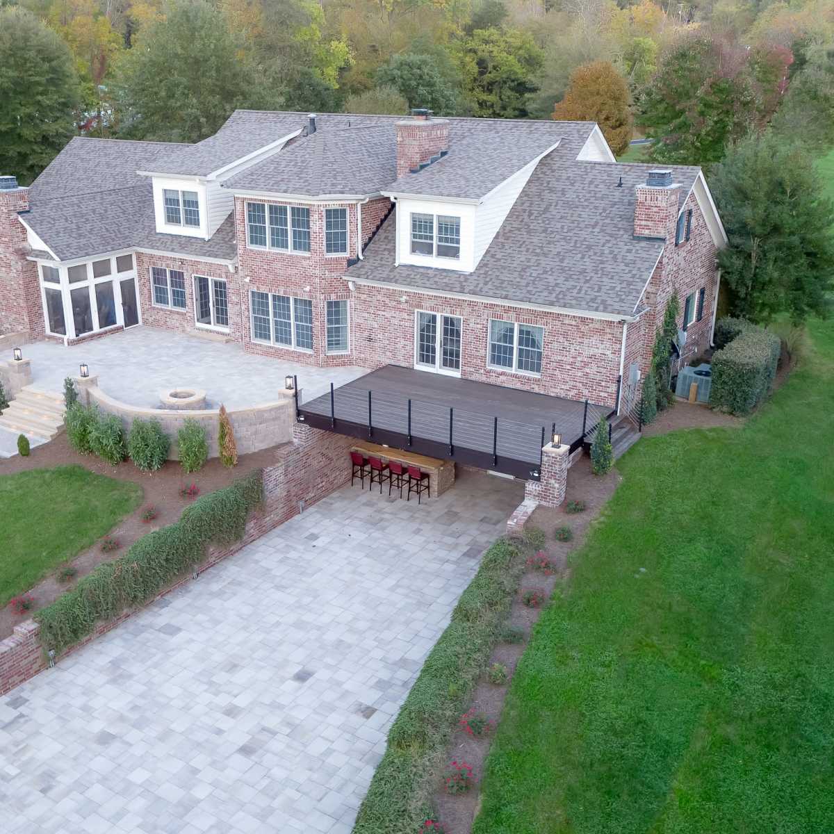 A large brick house with multiple gabled roofs, a stone patio, and a shaded outdoor dining area surrounded by greenery.