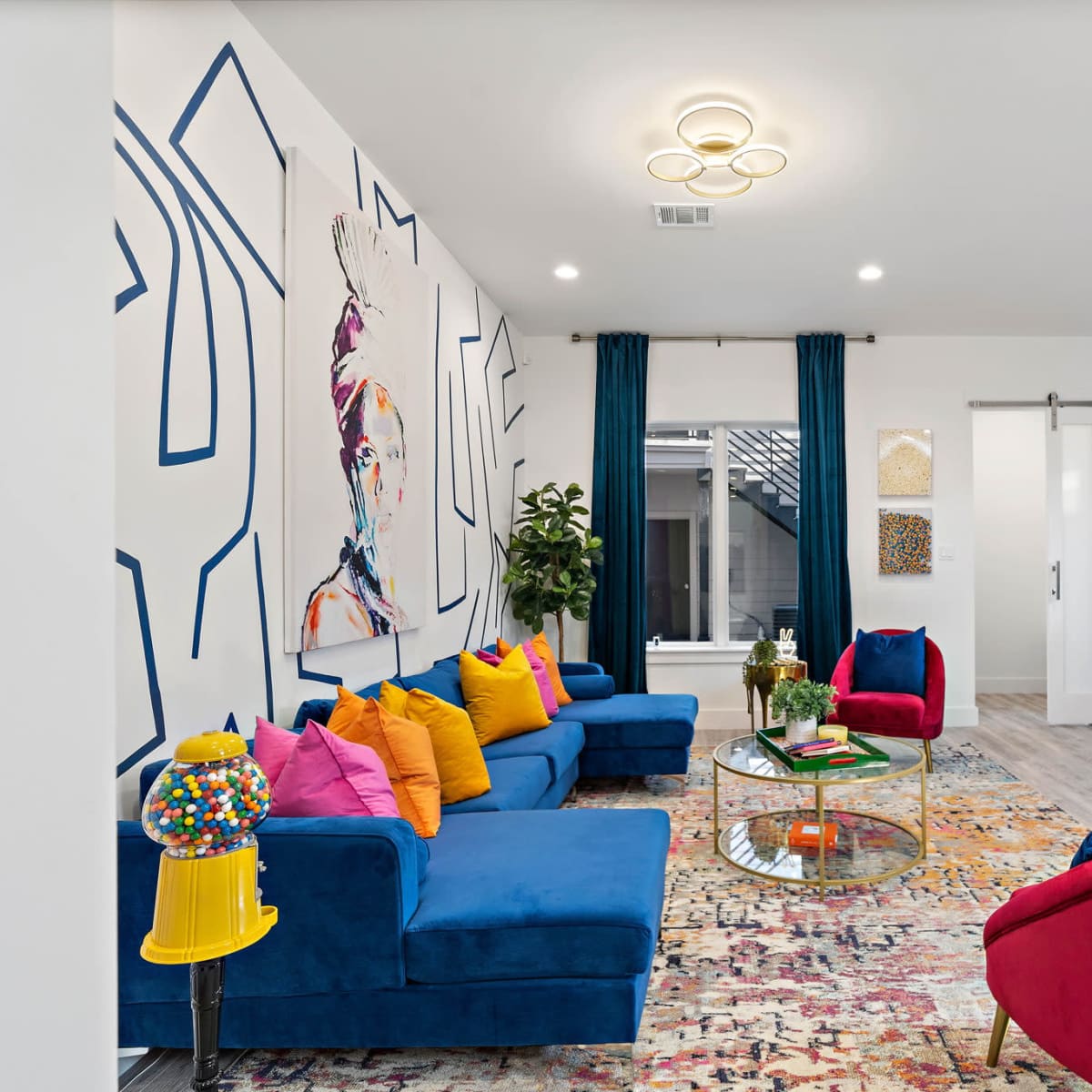 The room features a vibrant seating area with blue sofas, colorful pillows, and a round glass coffee table, surrounded by large artwork and decorative elements.