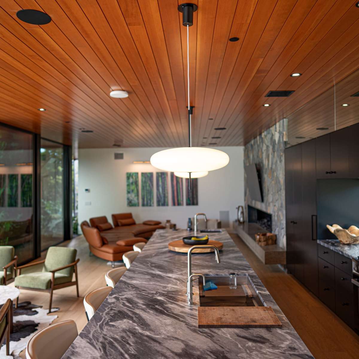 A modern kitchen with a marble countertop, wooden ceiling, and a cozy living area in the background featuring brown sofas and green chairs.