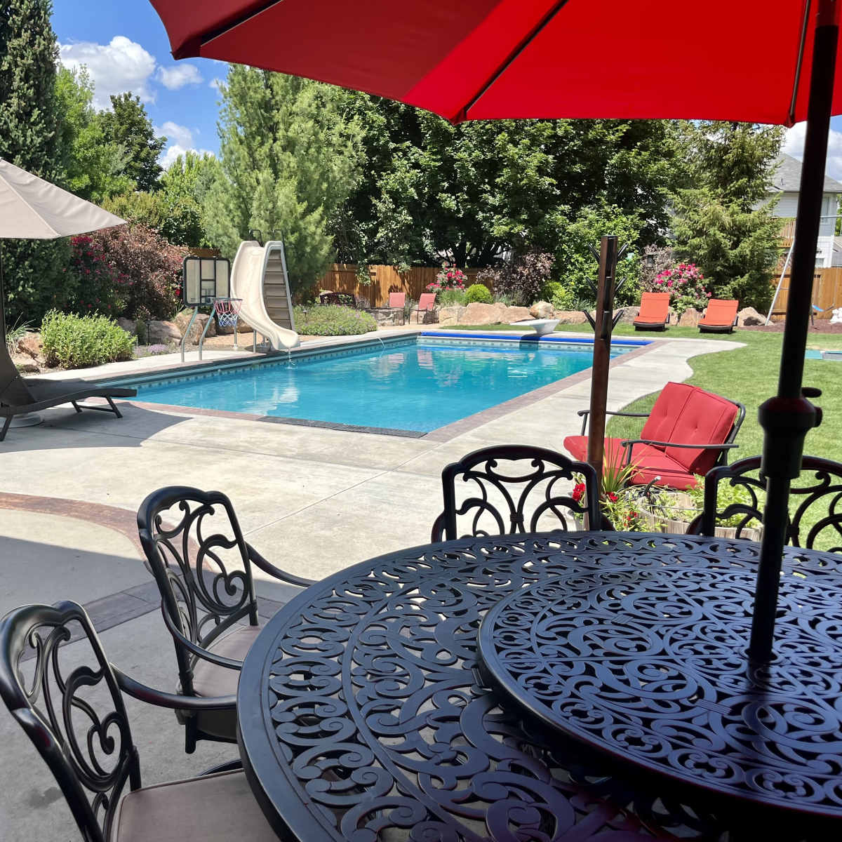 The scene shows a backyard pool area with a swimming pool, slide, lounge chairs, a dining table, and shade structures surrounded by landscaped greenery.