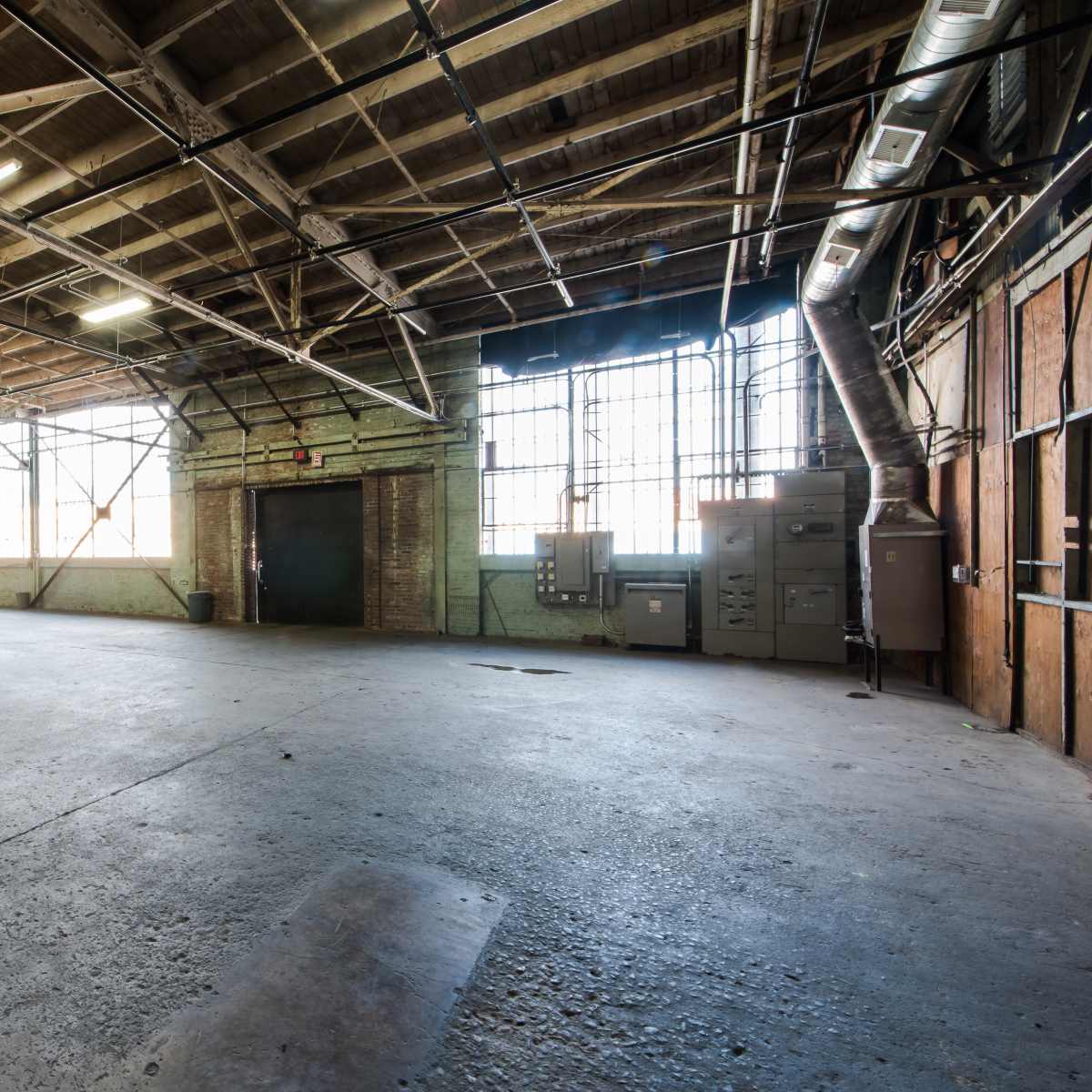 A large, empty industrial space with concrete flooring, exposed wooden beams, and large windows along one wall.