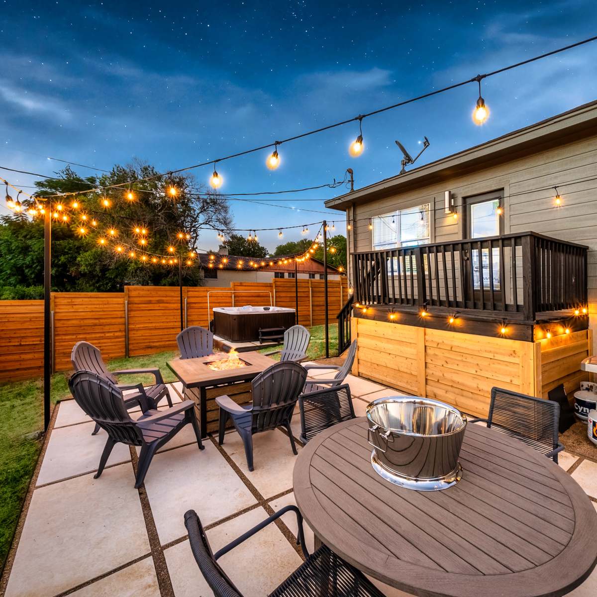 A backyard patio with string lights, a dining table, lounge chairs, and a barbecue grill beside a wooden fence.