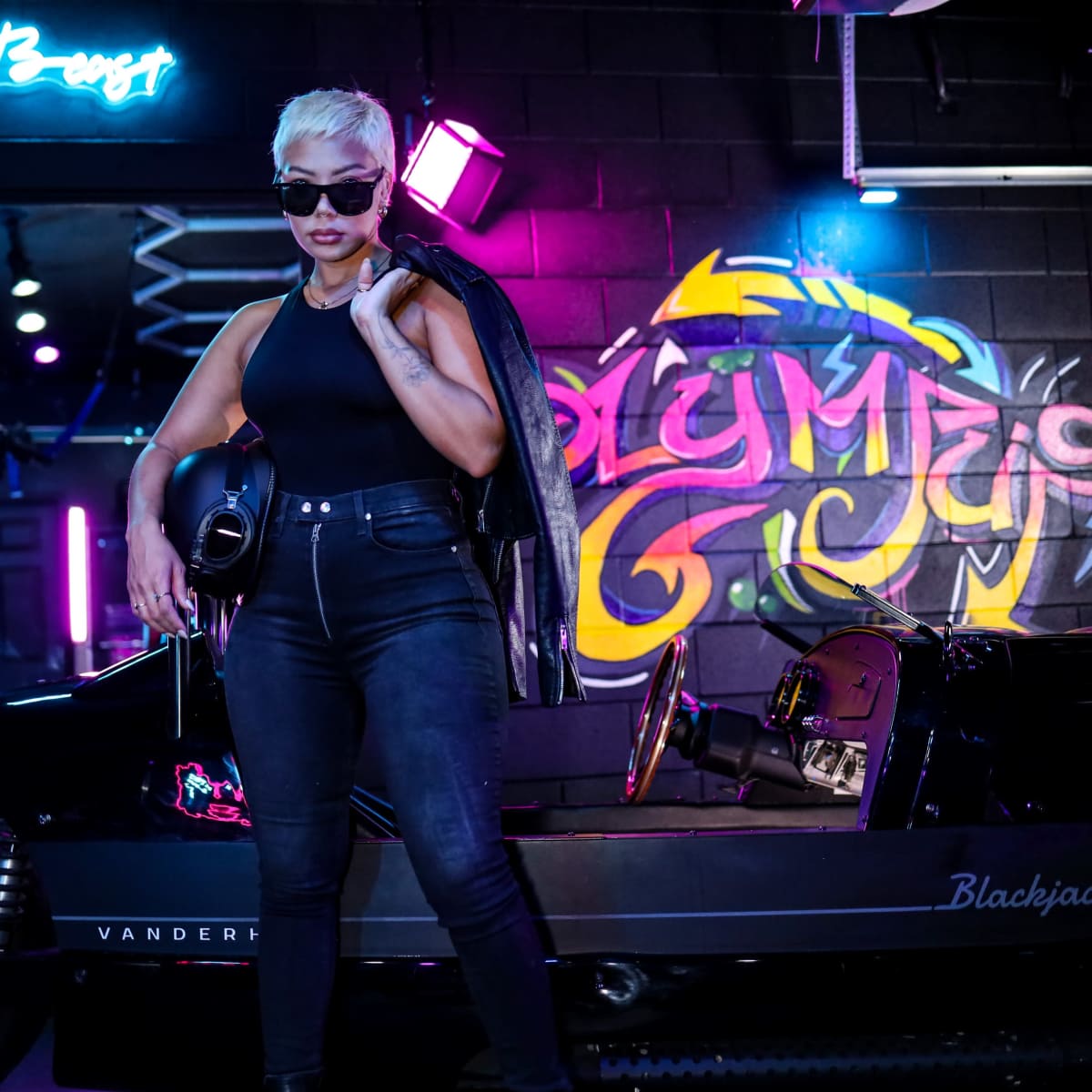 A person with short blonde hair and sunglasses stands beside a black vintage car in a colorful urban setting.