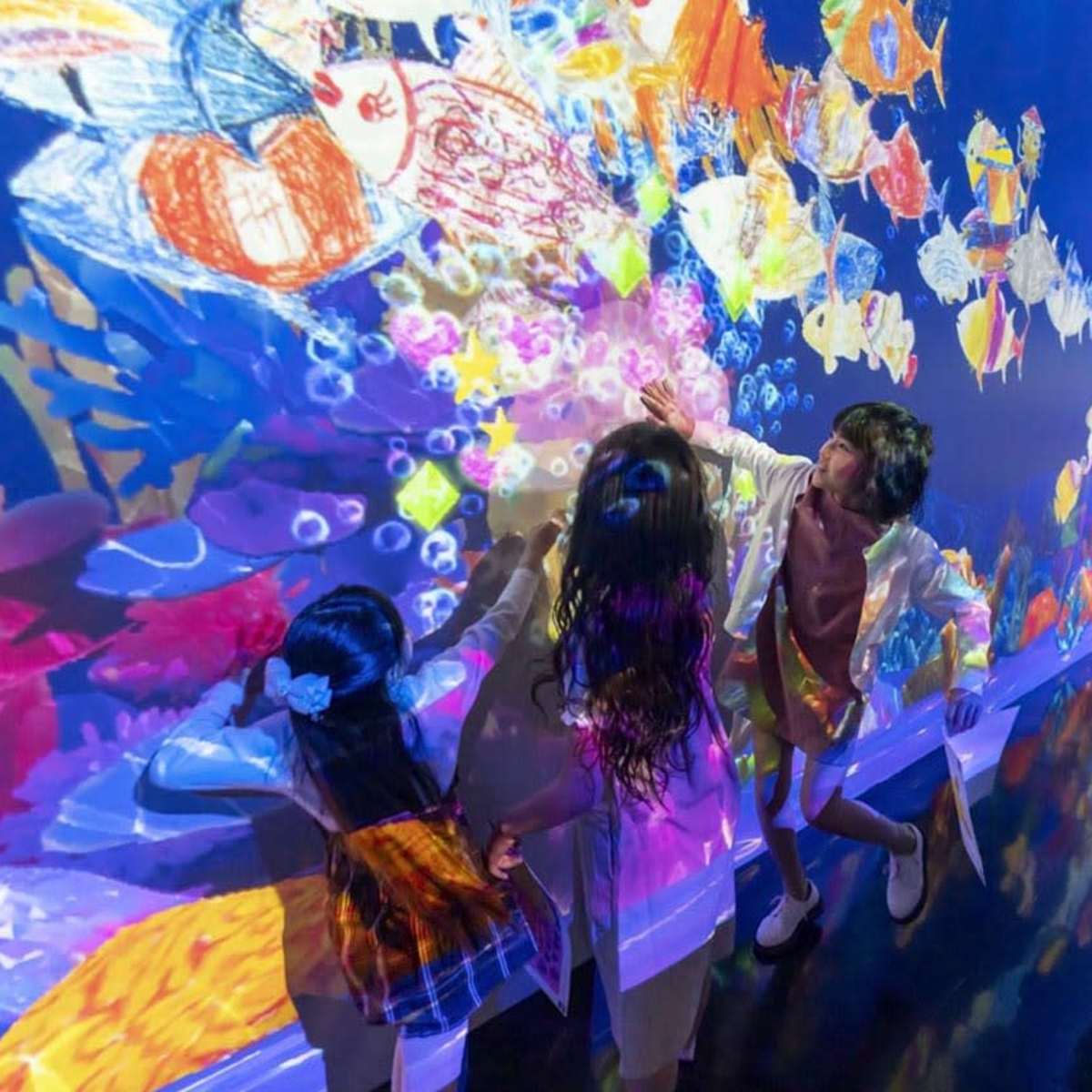 A group of children and an adult interact with a vibrant, projected underwater scene covered in colorful fish and bubbles.