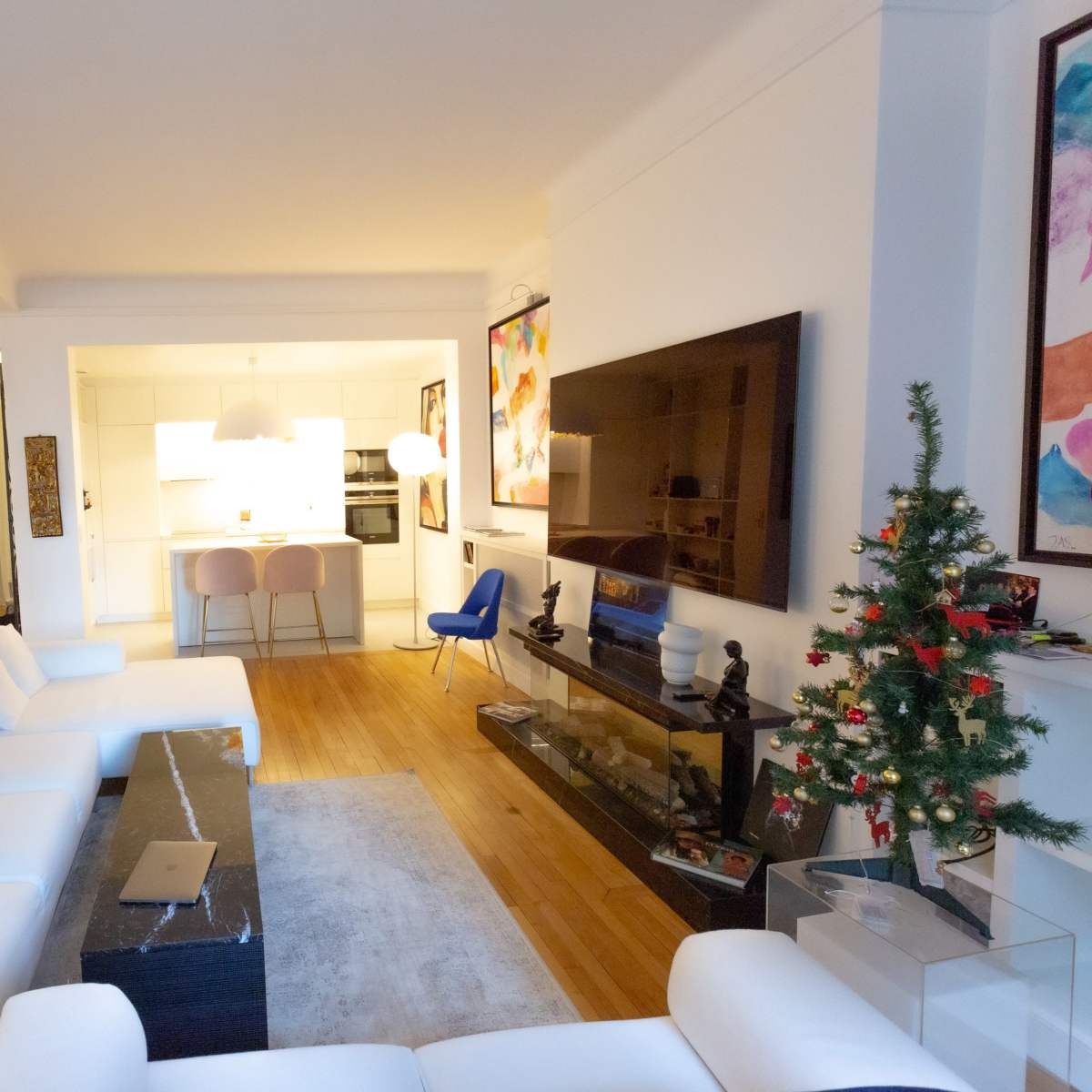 A modern living room with a white sectional sofa, a glass coffee table, a small Christmas tree, and an open kitchen in the background.
