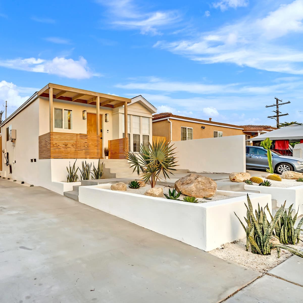 A modern single-story house with a wooden front porch is situated on a concrete driveway surrounded by landscaping featuring rocks and succulents.
