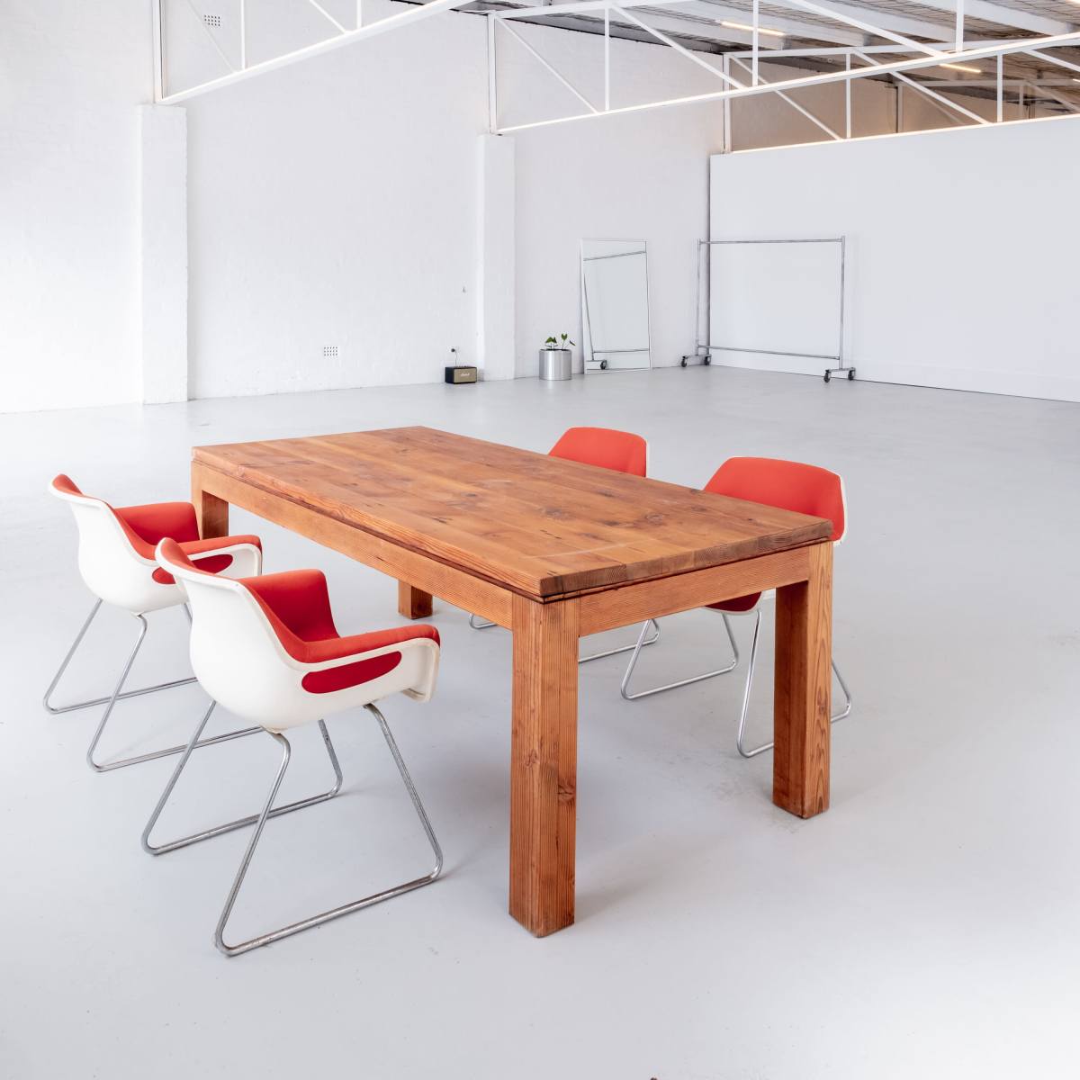 A wooden table with four chairs, two white and two red, is set in a spacious, empty room with white walls and a polished floor.