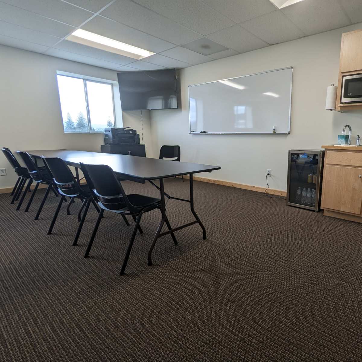A conference room with a long table, black chairs, a whiteboard, a microwave, a small refrigerator, and trash bin against a wall.