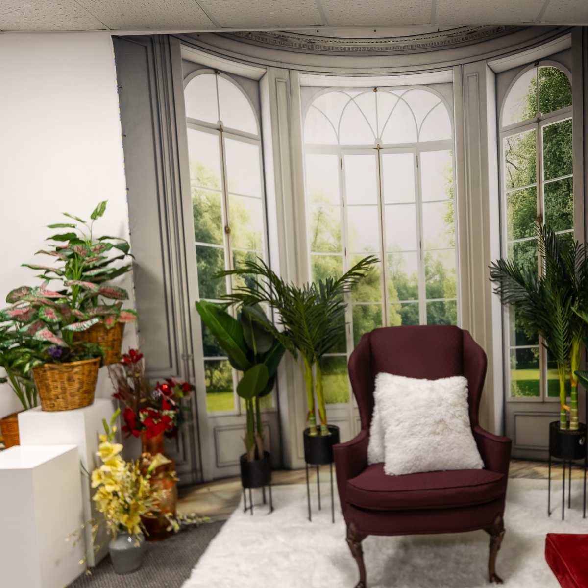 A cozy seating area with a burgundy armchair and a red sofa, surrounded by potted plants against a backdrop of large windows.