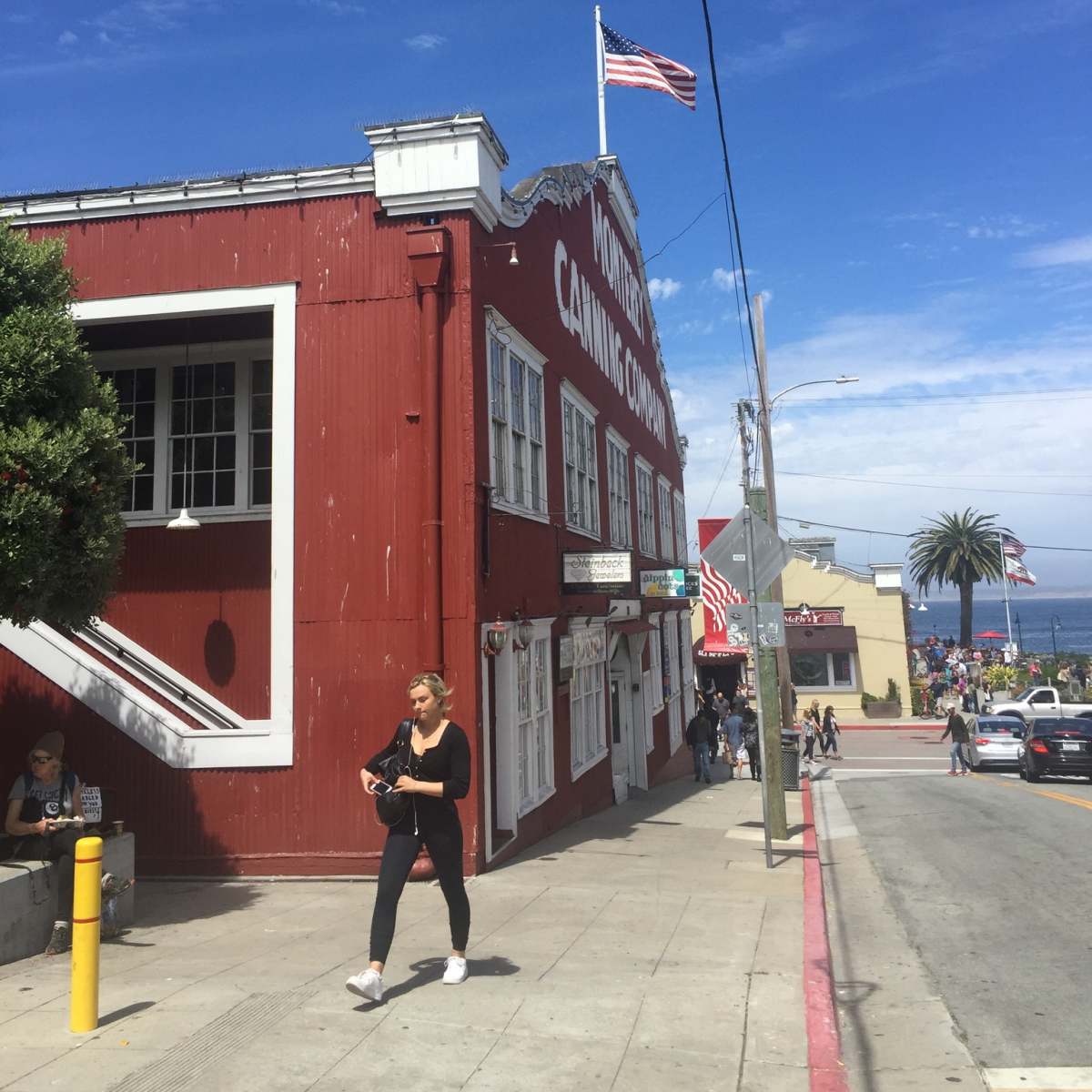 A woman walks past a red building marked "Monterey Canning Company" with an American flag flying above, while the ocean is visible in the background.