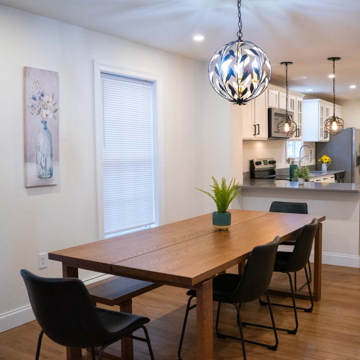 A modern dining area with a wooden table, four black chairs, and a kitchen visible in the background, featuring light cabinets and pendant lighting.
