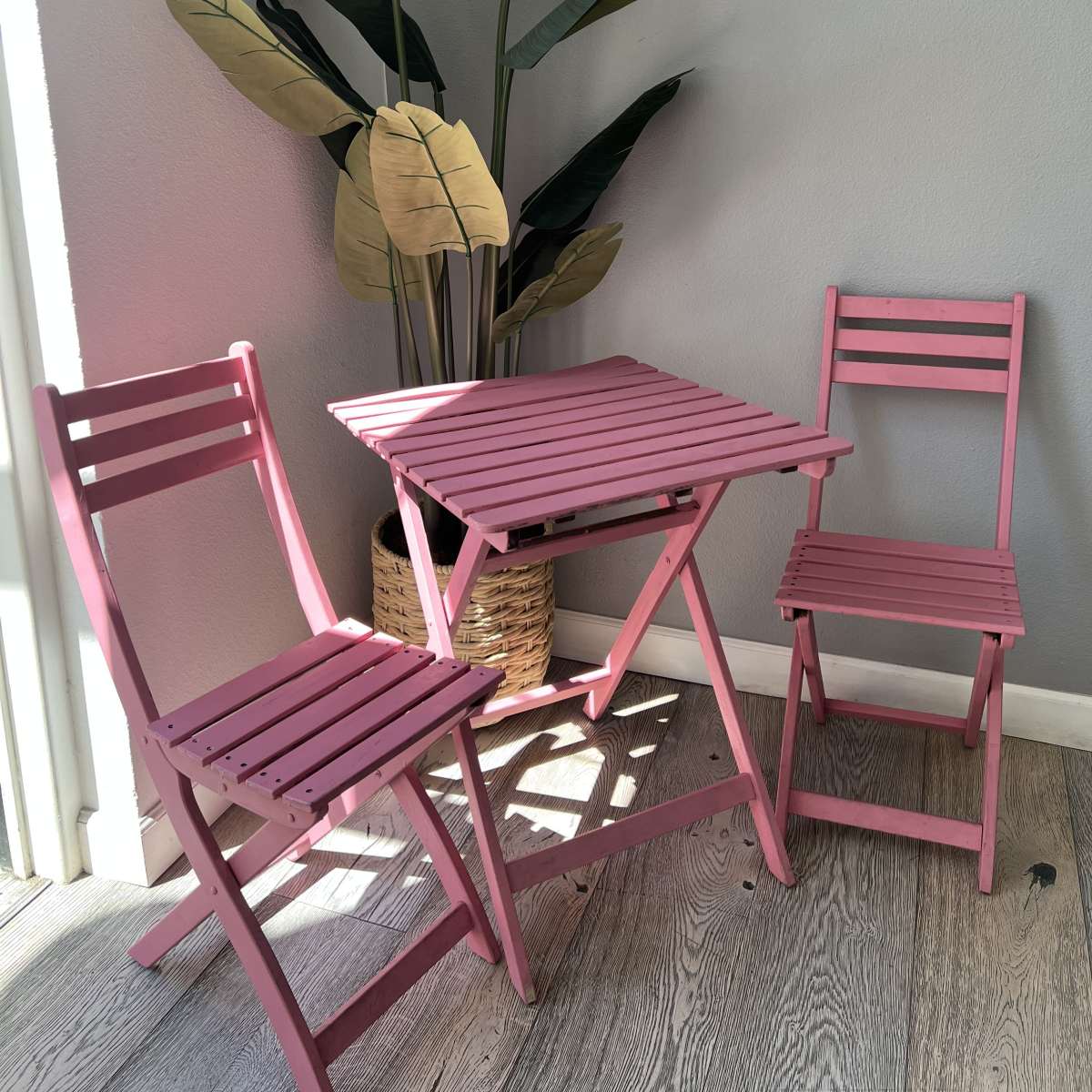 A small pink table and two matching chairs are positioned next to a large leafy plant in a well-lit corner.