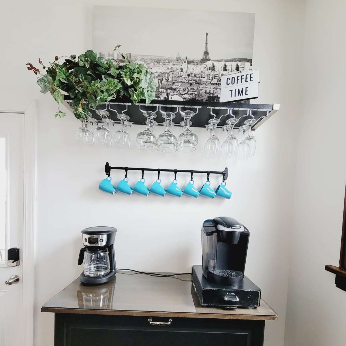 A coffee station with a black countertop, a coffee maker, a Keurig machine, a shelf holding glasses, and blue mugs, along with a decorative plant and a sign that reads "COFFEE TIME."