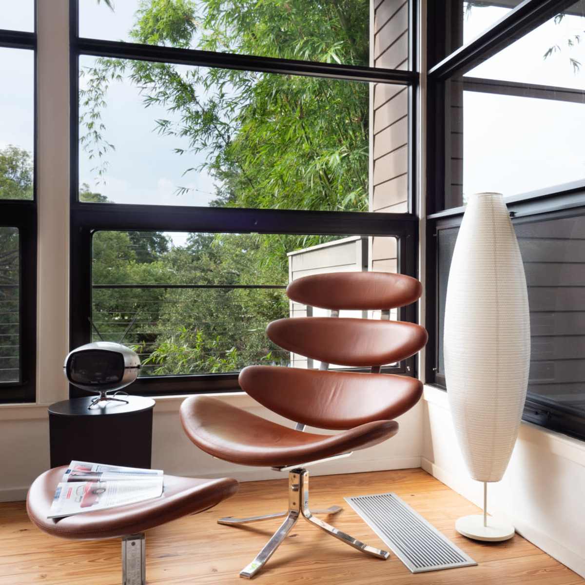 A modern seating area features a brown, curved chair and matching ottoman next to a tall, white floor lamp, with large windows revealing greenery outside.
