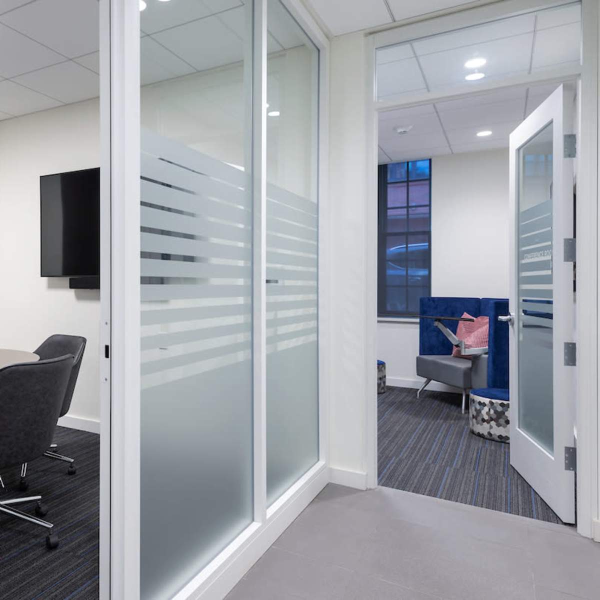 A modern office space with a conference room featuring a round table and chairs, adjacent to a lounge area visible through an open door.