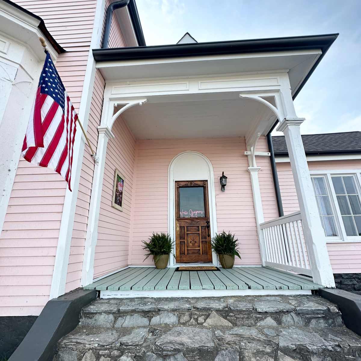 A pink house with a front porch, featuring a wooden door and an American flag hanging to the left.